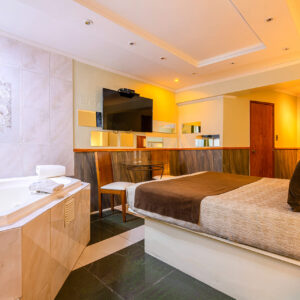 Habitación luminosa con cama doble, jacuzzi en el costado y decoración en tonos cálidos, equipada con televisor de pantalla plana y un pequeño set de sillas y mesa.